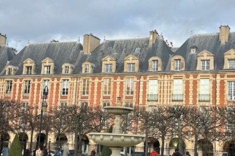 Park with fountain, people walking, and historic building in background under cloudy sky.
