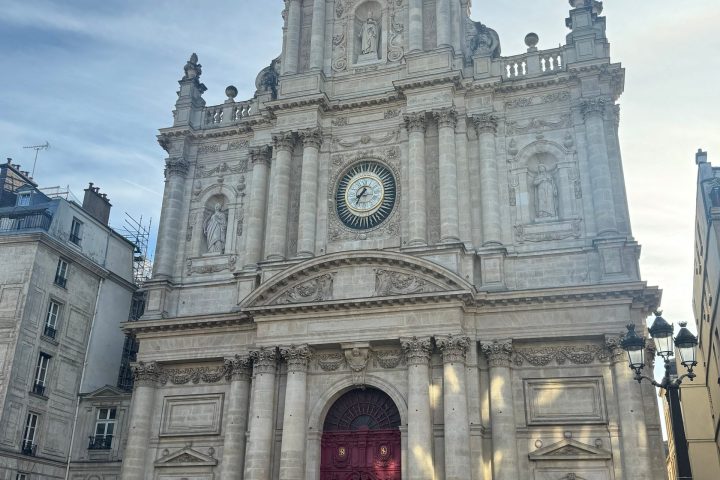 Baroque-style church facade with red doors, clock, and statues on a clear day.