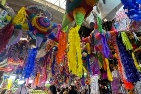 Colorful piñatas hanging in a lively market with shoppers and various stalls.