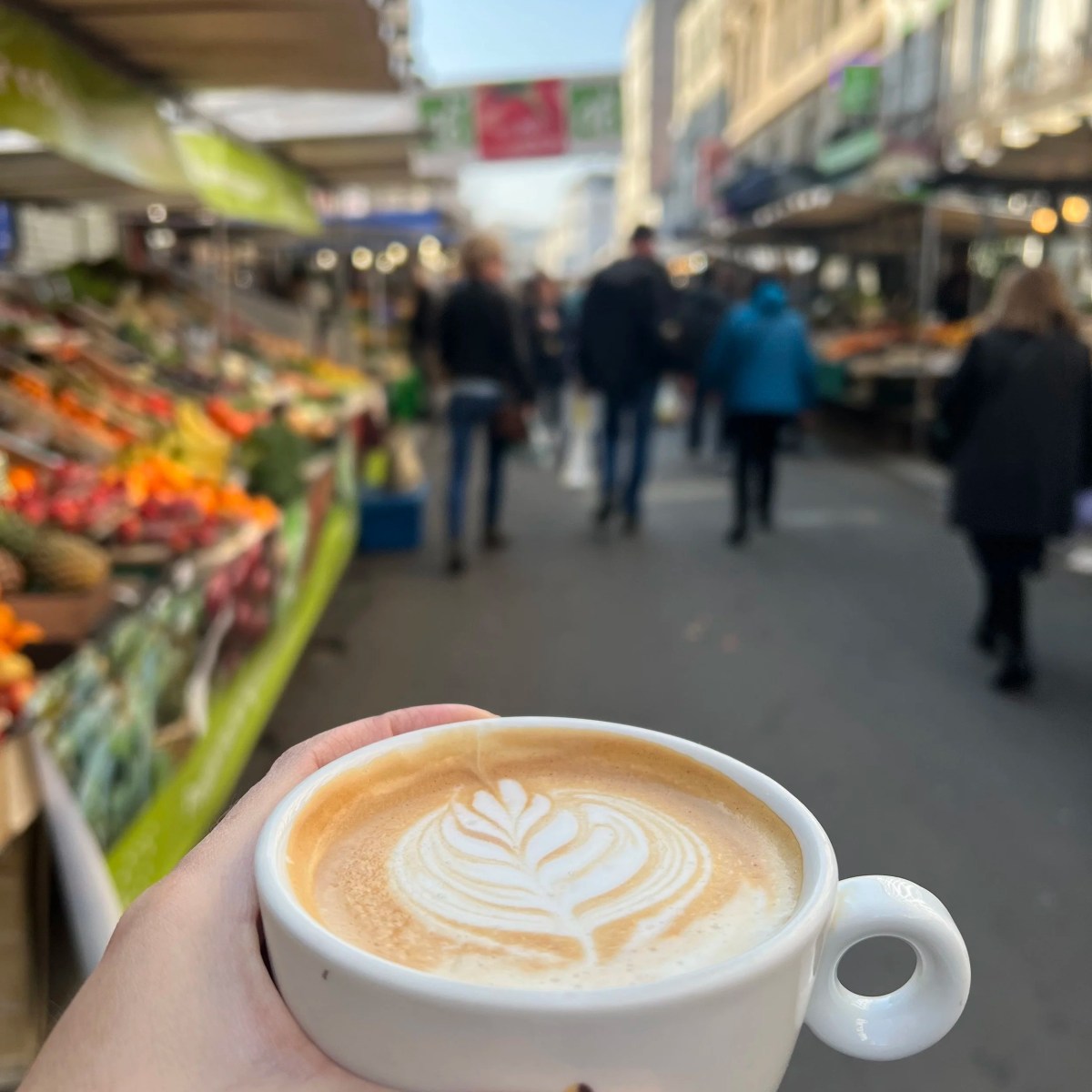 Hand holding latte with frothy art at an outdoor market.